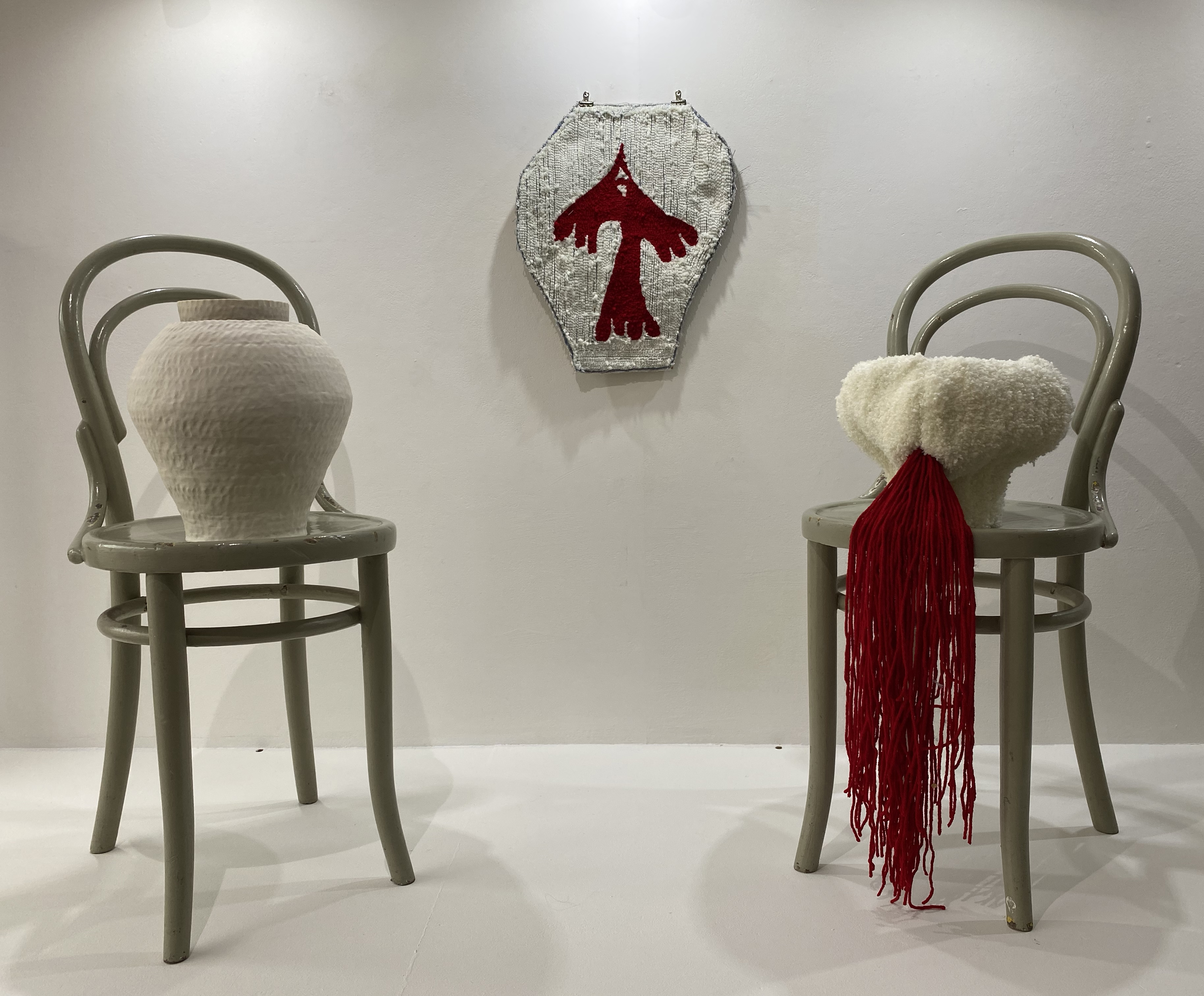 Two white vessles in conversation on soft green bentwood chairs. One is ceramic, the other is rugged. On the wall in between the chairs is a third white vessle showing a red bird inside. Long bits of red yarn spills out of the rugged vessle.