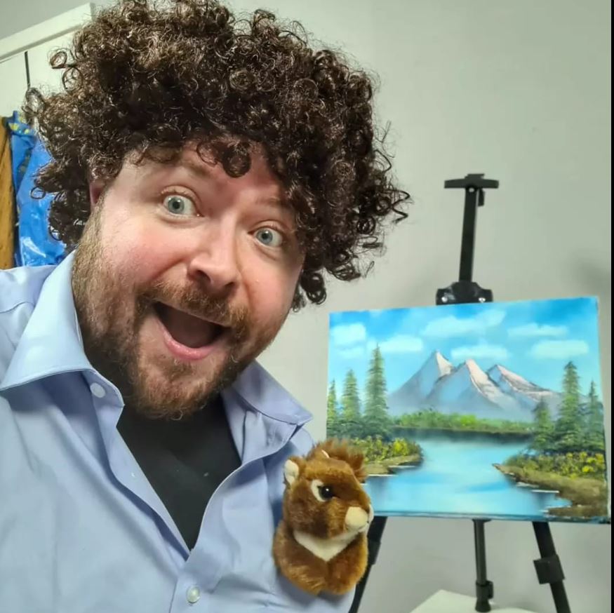 Workshop attendee smiling while wearing a Bob Ross wig, with a toy squirrel in their top pocket, beside their Bob Ross landscape painting on an easel