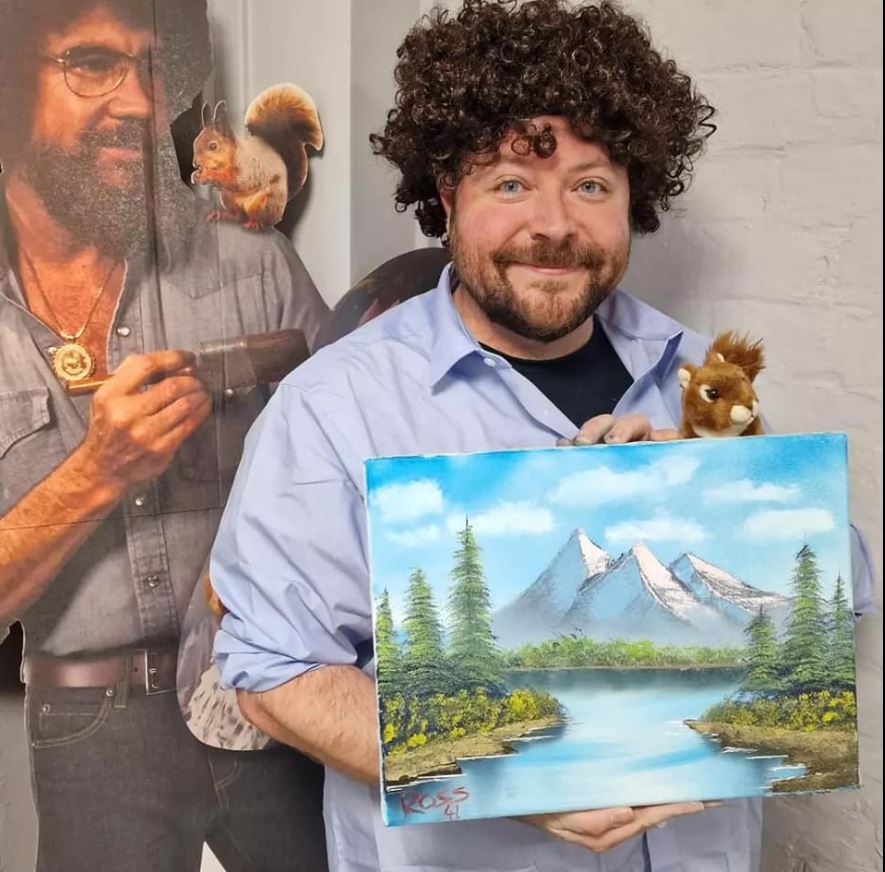 Workshop attendee proudly holds his painting next to a life size cut out of Bob Ross